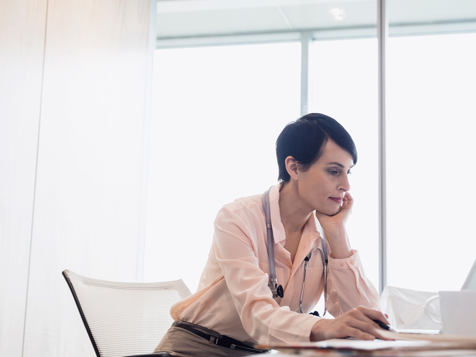 A doctor in a white coat sits at a desk, focused on a laptop, in a bright office. Stethoscope around neck; conveys concentration and professionalism.