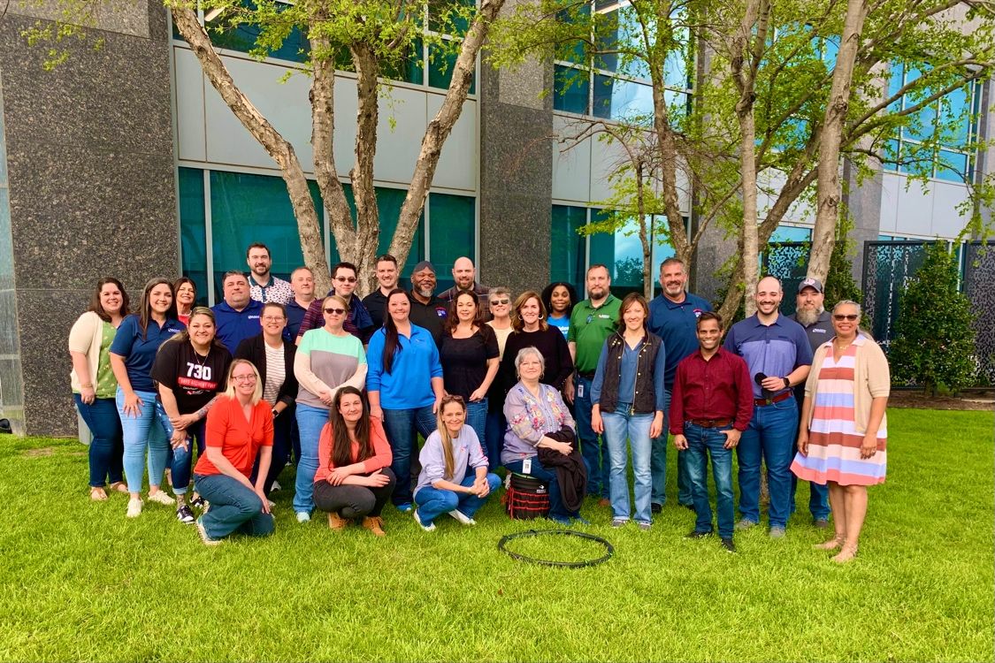 A large group of people posing for a group photo outside, standing and sitting on a grassy area in front of a modern office building with glass windows. Trees frame the scene, and the participants are smiling, suggesting a team bonding or corporate event.