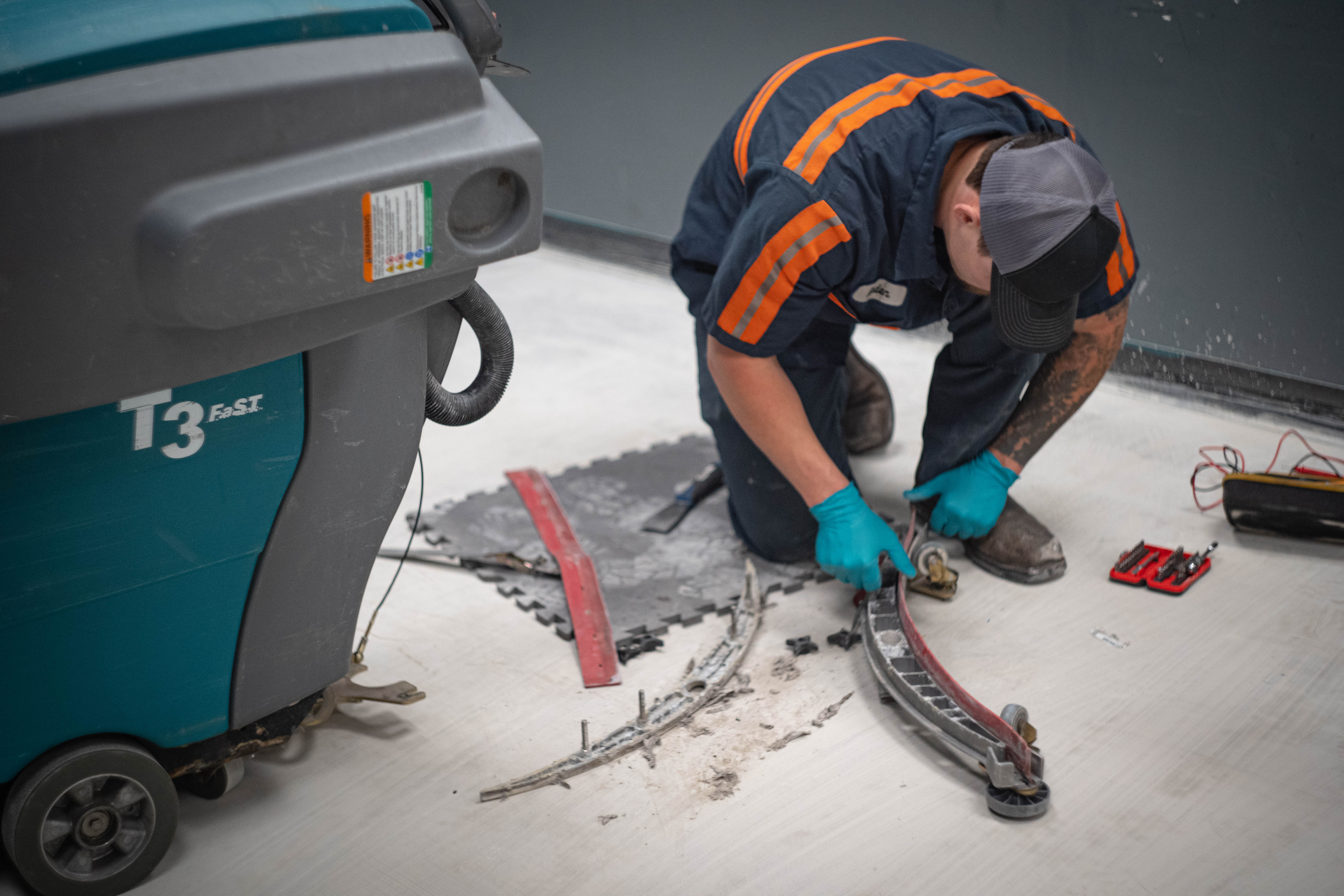 A WaySolutions field technician replaces the heavy-duty squeegee blades and services the aluminum pickup tool assembly for a teal Tennant T3 FAST walk-behind floor scrubber. Kneeling on a service mat with professional tools spread out, the technician fits precision original parts to guarantee streak-free water recovery for demanding commercial environments like retail grocery stores.