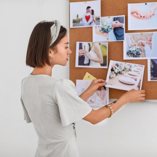A woman with short brown hair and a headband is pinning up photos on a corkboard. The photos appear to be wedding-themed, featuring couples, wedding rings, and floral arrangements. She is wearing a light-colored dress and jewelry.