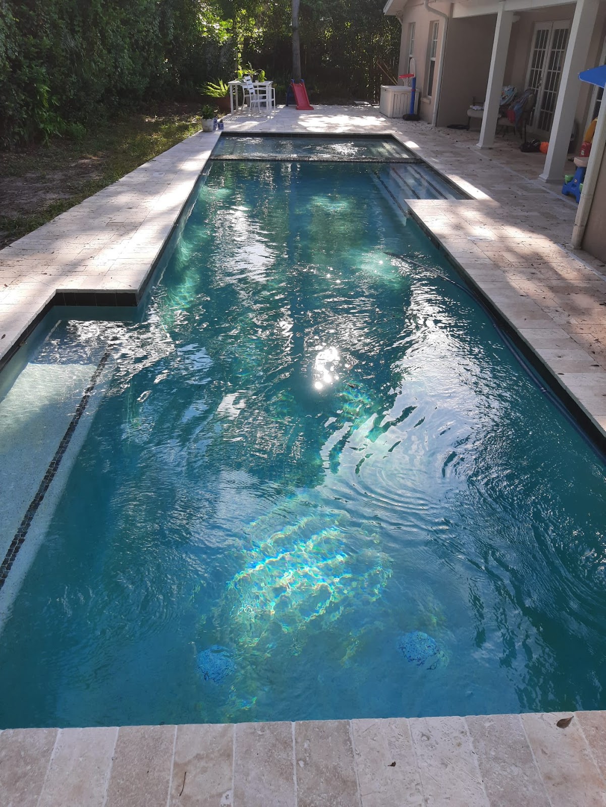 Long narrow residential swimming pool with deep blue water, light stone pavers, and a shaded backyard setting.