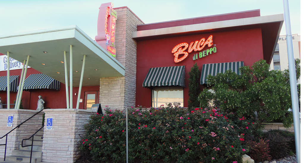 Buca di Beppo Austin signs with greenery bushes, flowers, and green and white striped windows.