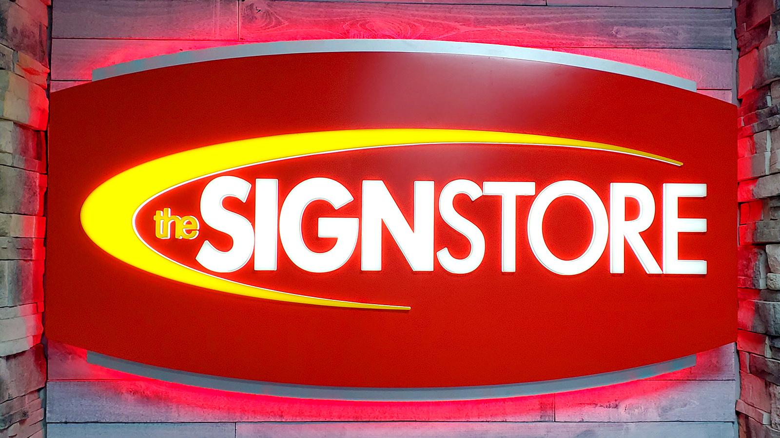 The image shows an illuminated sign for "The Sign Store" on a stone wall. It has a red background, bold white letters, and a yellow swoosh around the text. The sign is backlit, creating a warm glow that highlights its contours, making it stand out.