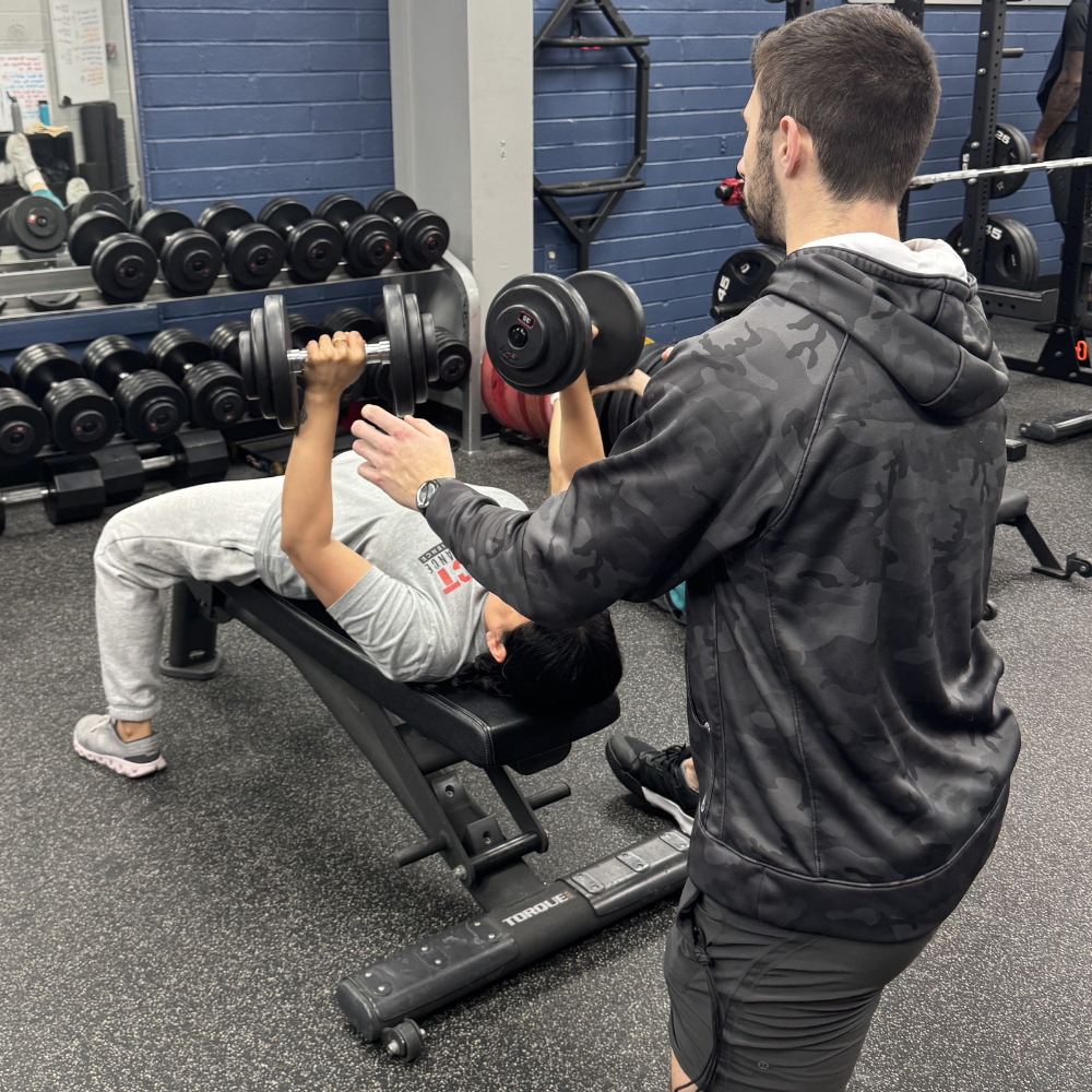Coach spotting female athlete during dumbbell bench press at Impact Sports Performance