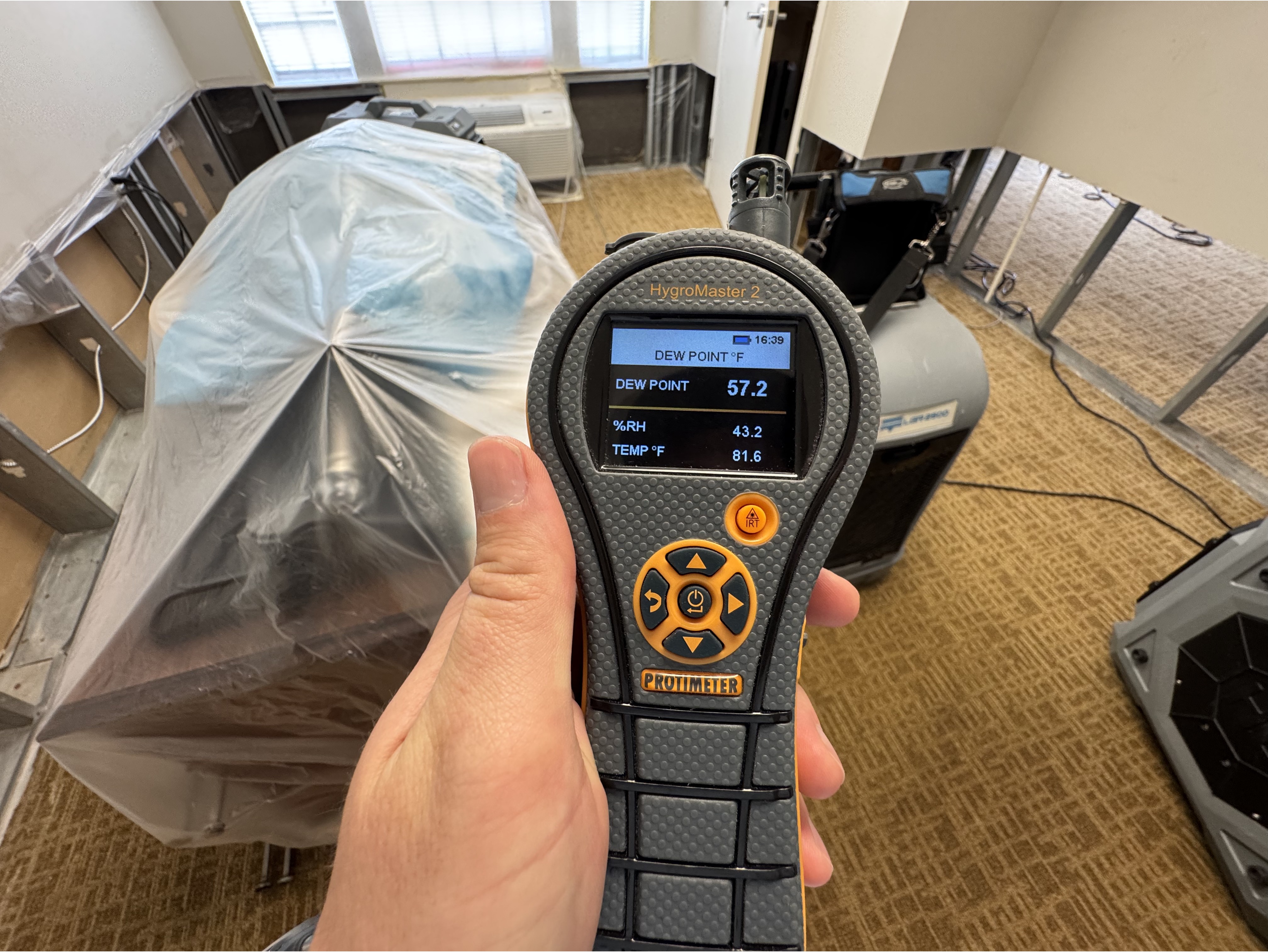 A hand holds a digital hygrometer displaying dew point, humidity, and temperature. The background shows a room with covered furniture and exposed walls.