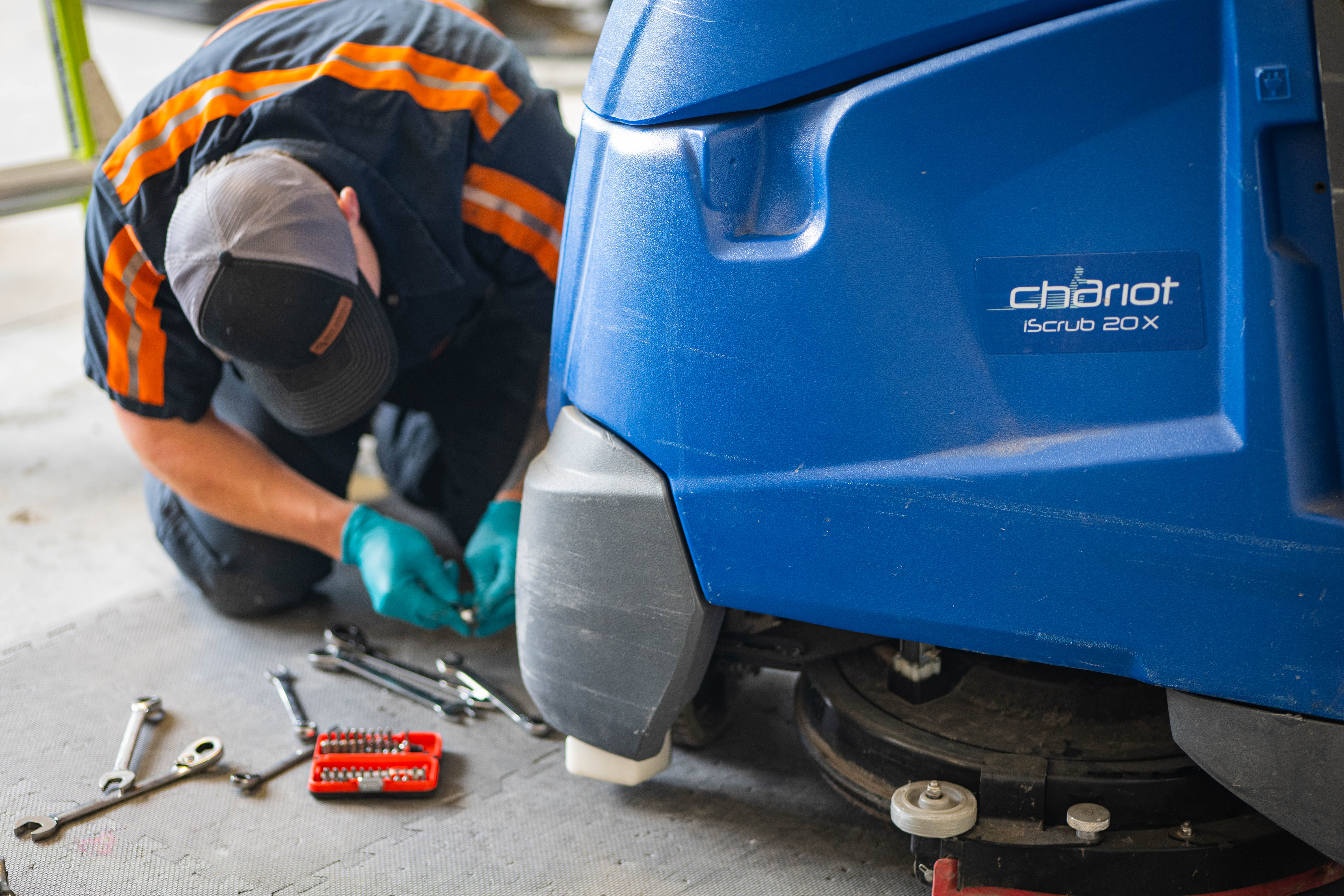 A WaySolutions technician kneels on a protective service mat to perform ground-level mechanical repairs on a blue Kärcher Chariot iScrub 20X stand-on floor scrubber. Serving commercial facilities in Layton, Utah, our experts use professional hand tools to thoroughly maintain and tighten essential undercarriage hardware.
