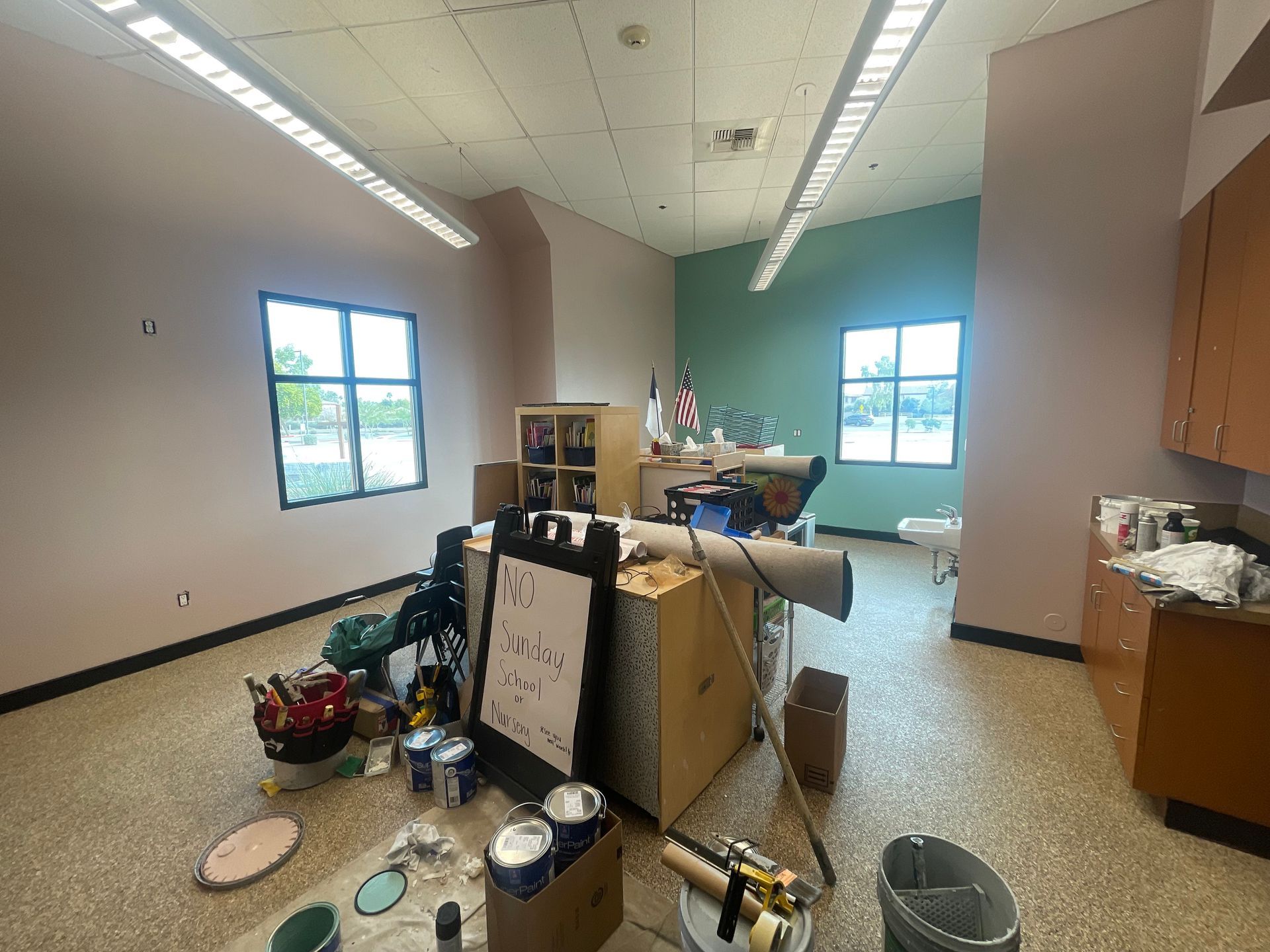 A classroom or workspace under renovation with painting supplies, tools, and furniture scattered around, featuring large windows, cabinets, and bright lighting, indicating ongoing interior improvement and maintenance work.