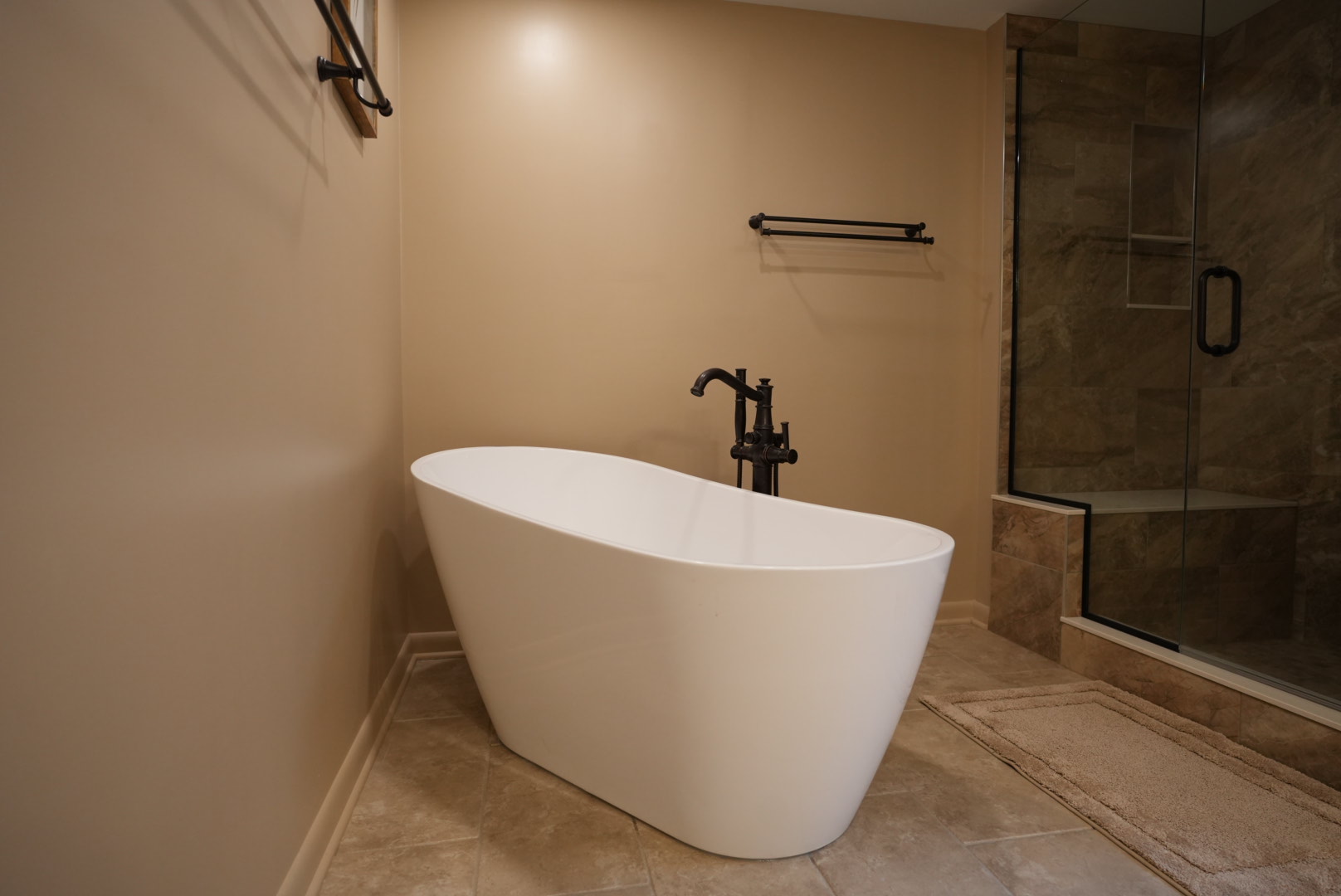 This bathroom remodel features a modern freestanding soaking tub paired with a sleek floor-mounted faucet, a custom tile walk-in shower with frameless glass shower doors, and warm-tone tile flooring.