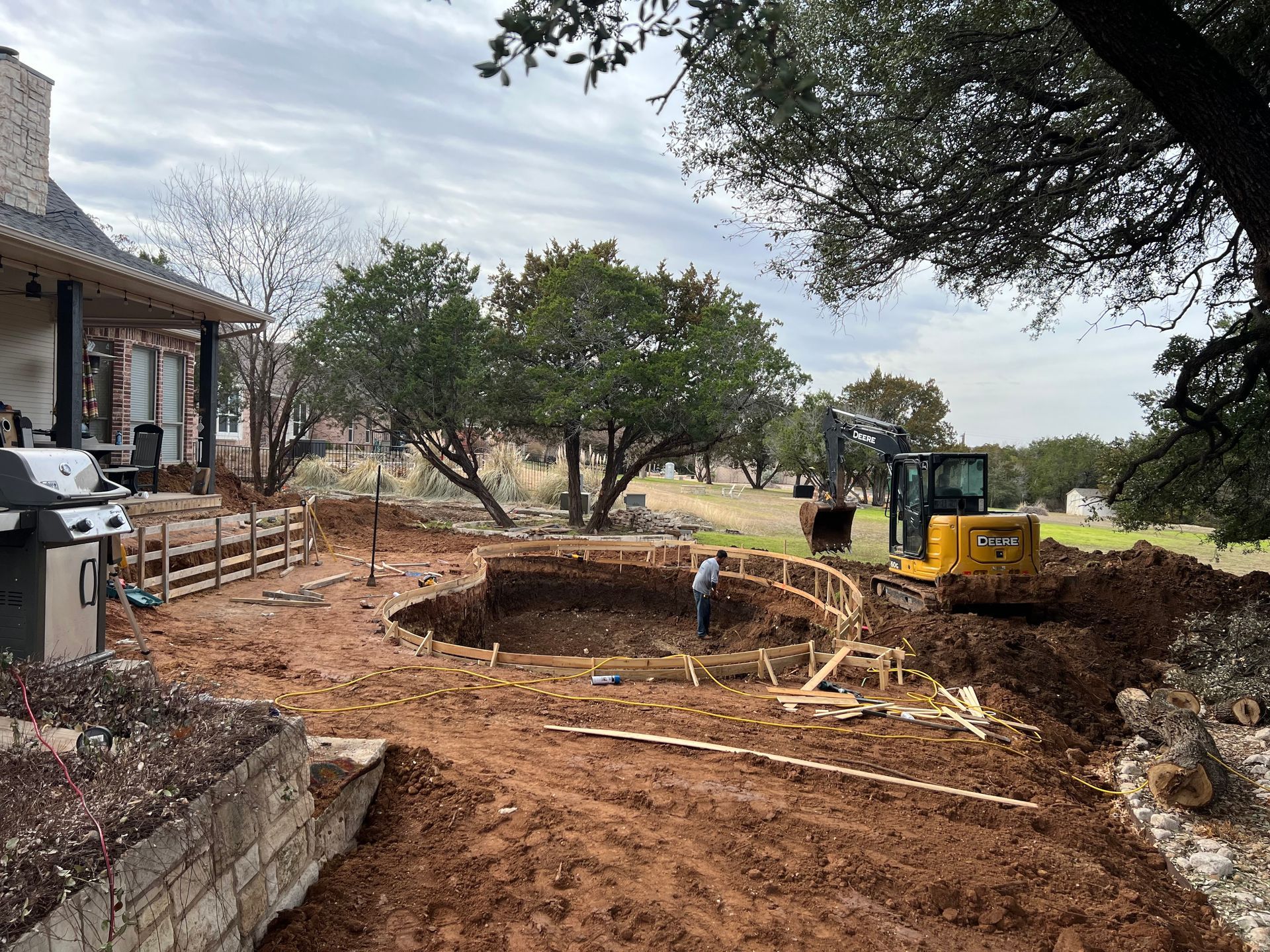 A worker stands inside a newly excavated pool pit while a John Deere excavator moves dirt in the background. Wooden forms outline the pool's curved shape near a brick house.