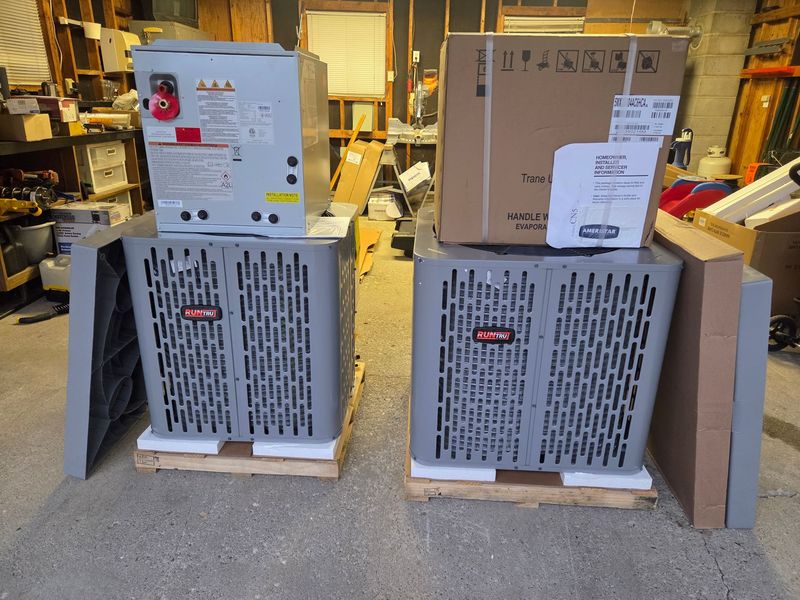 Two brand-new RunTru by Trane air conditioning condensers sit on wooden pallets in a workshop, topped with boxed components and installation manuals, ready for a professional residential HVAC installation.
