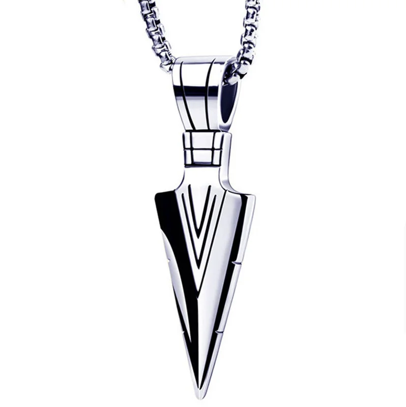 A close-up studio shot of a silver-colored arrowhead pendant necklace against a white background. The pendant is designed with engraved lines that create a V-shape, and it is suspended from a thick, silver-colored chain with a box link pattern. The pendant is highly polished and reflects light.
