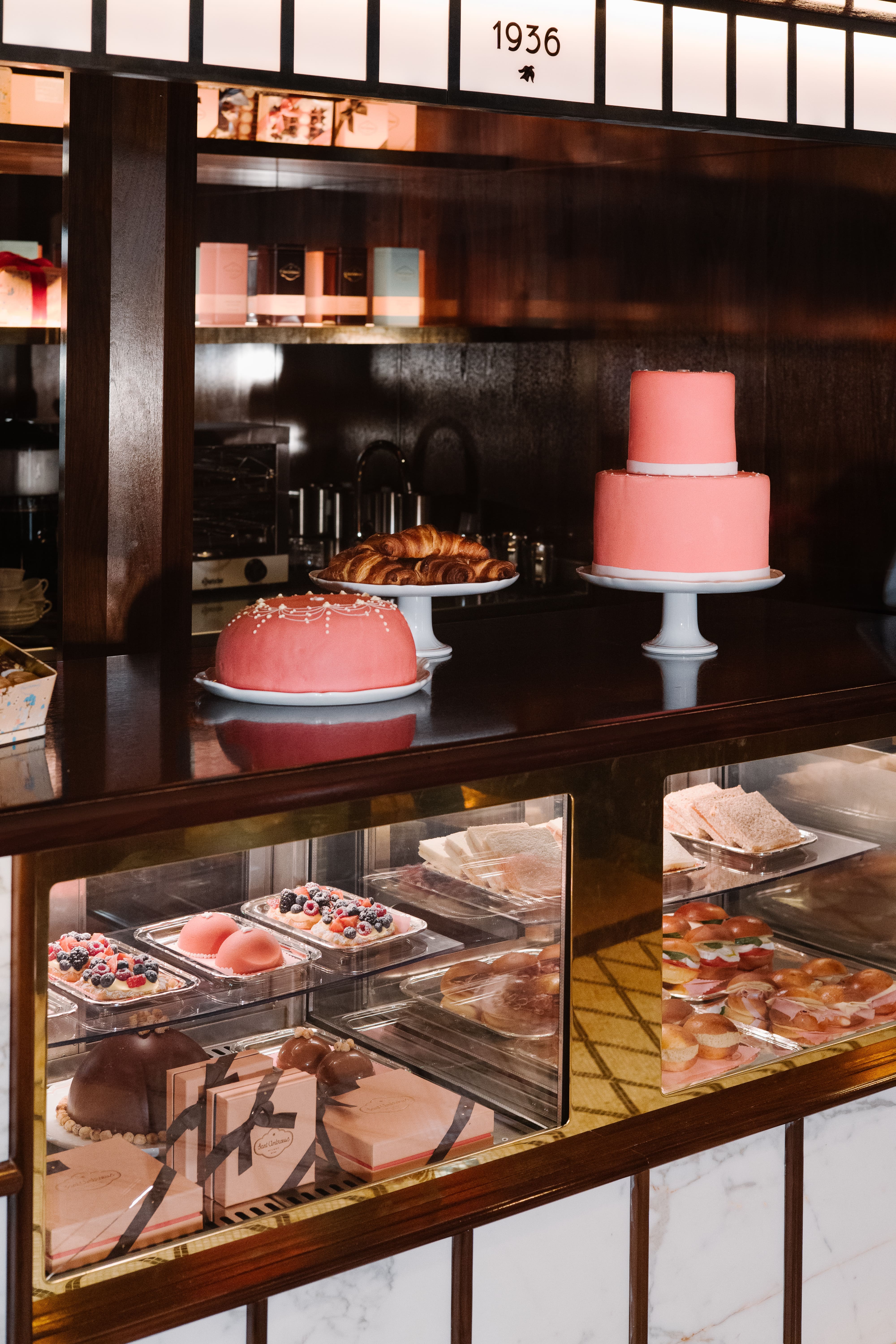 Pastry display inside a classic café, featuring pink frosted cakes on pedestal stands, croissants, assorted pastries and sandwiches behind glass, with dark wood cabinetry and a softly lit, elegant ambiance.