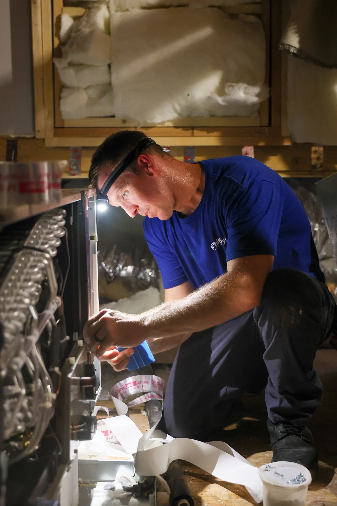 A man in a blue t-shirt and work pants is kneeling and working on what appears to be an HVAC unit in 