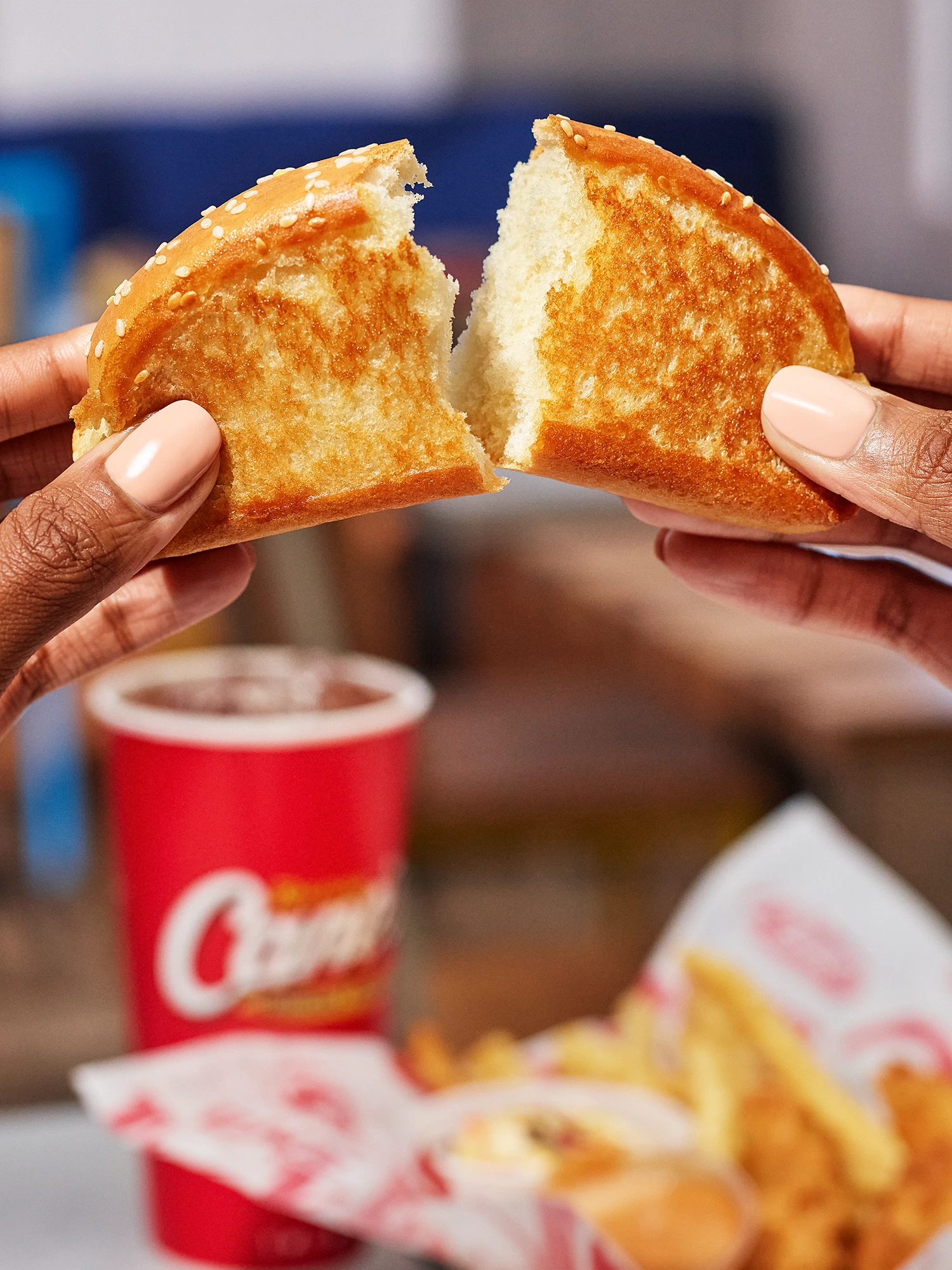 Two hands pulling apart a piece of toasted garlic bread (Texas toast), revealing the soft white bread inside, with a blurred meal and red cup in the background.