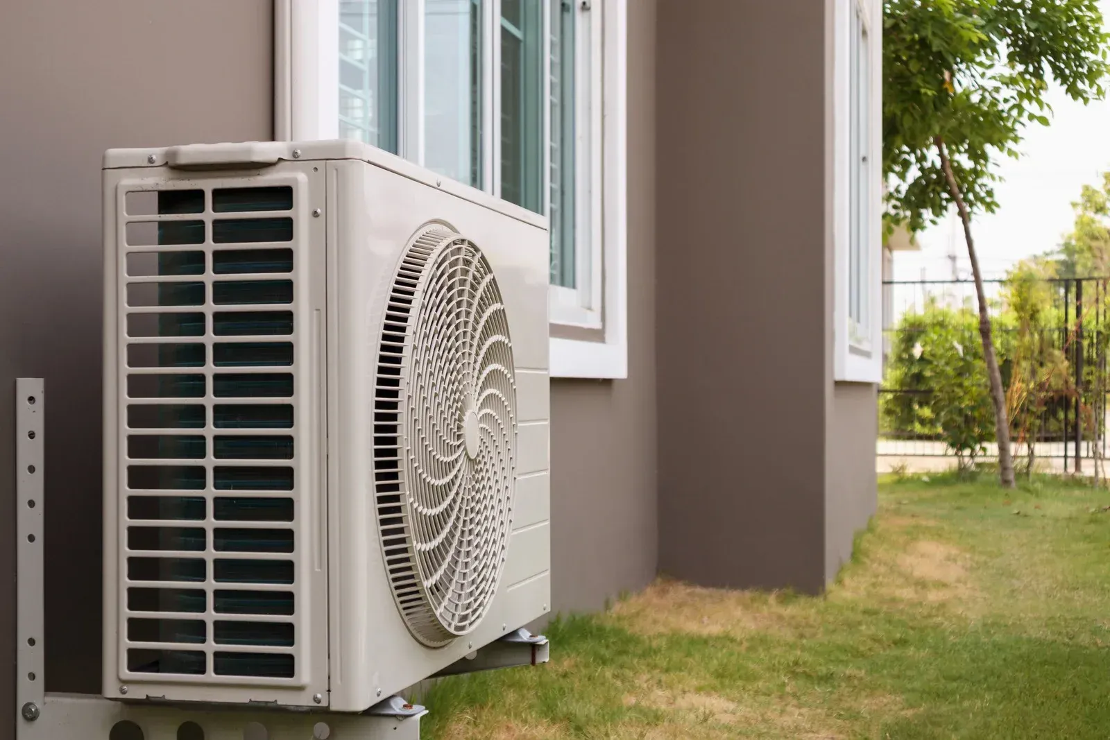 An outdoor air conditioner condenser mounted beside a house with windows, positioned slightly above ground on a metal stand, surrounded by grass and small trees in a residential backyard setting.