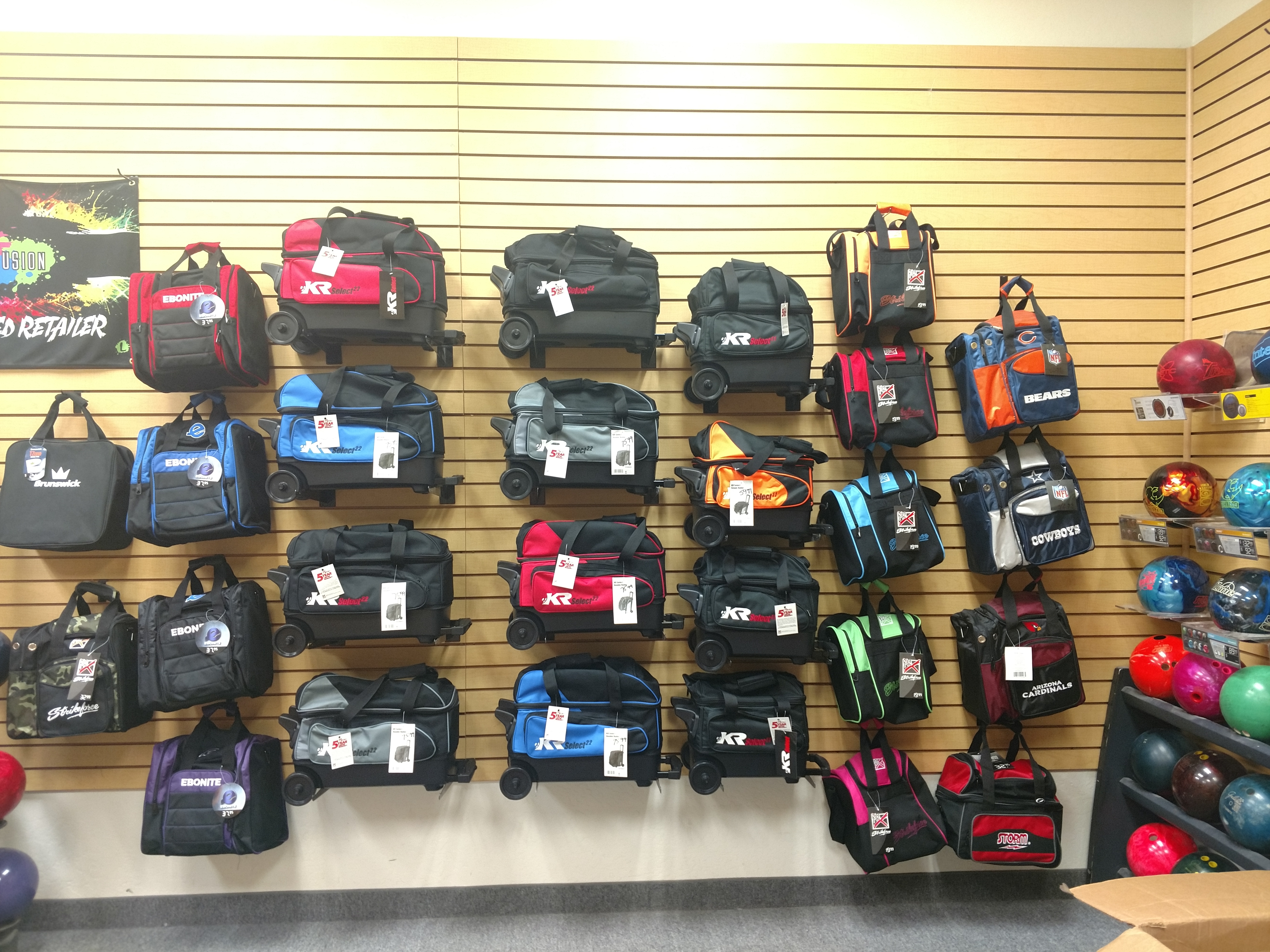 A store shelf displays a variety of bowling bags and bowling balls. The bowling bags are mostly black, with some accents of red, blue, green, and pink. Several bags have wheels and retractable handles, indicating they are designed for travel. The bowling balls are various colors including red, pink, green, blue, and black. Some bags feature brand logos such as Ebonite, Brunswick, KR Select, and Strikeforce. Team logos like the Chicago Bears and Dallas Cowboys are also visible on some bags. The items are arranged on a light wood-paneled wall with horizontal slats, typical of a retail store display.