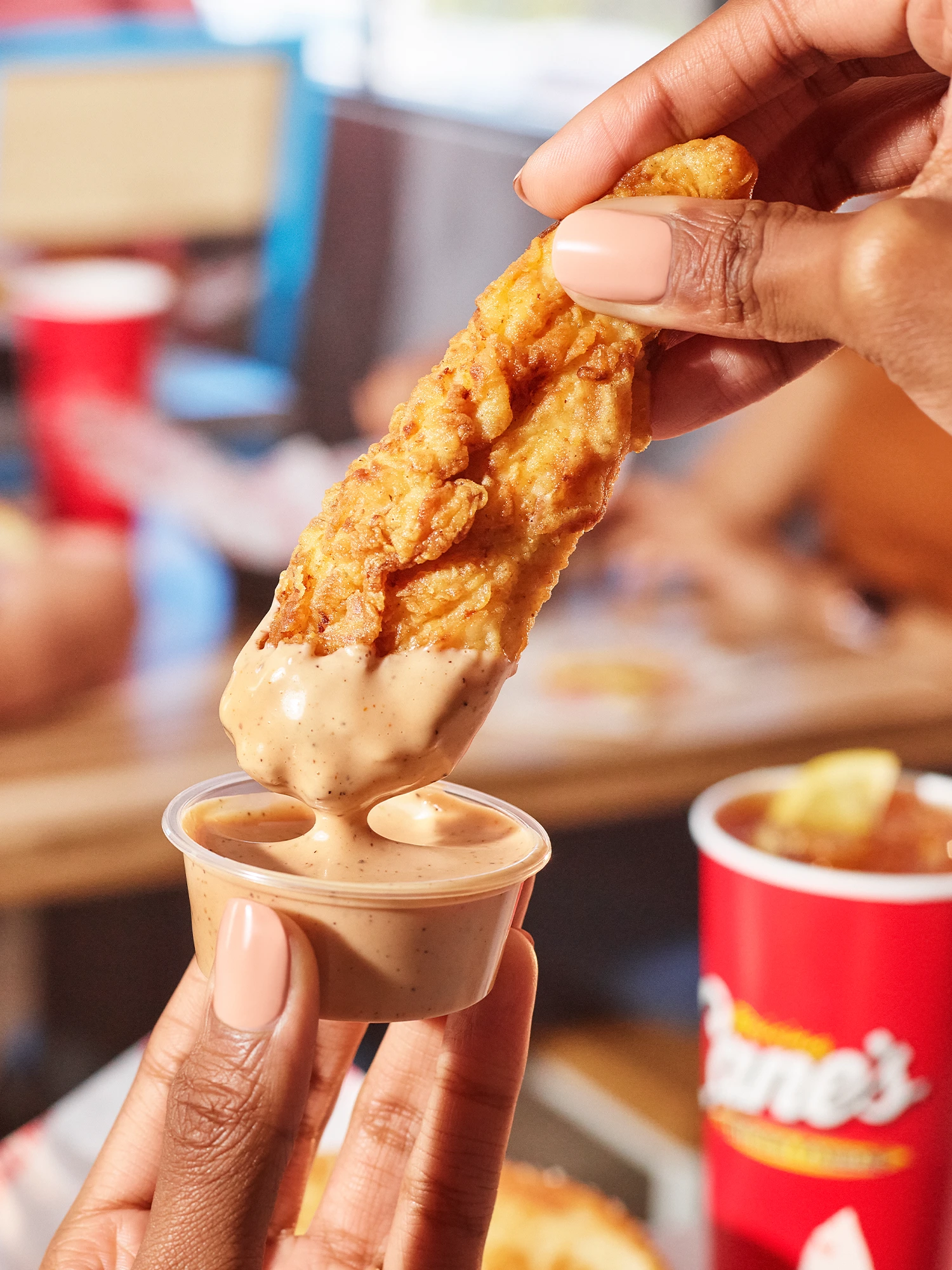A close-up action shot of a hand dipping a crispy chicken finger into a small cup of Cane's sauce.