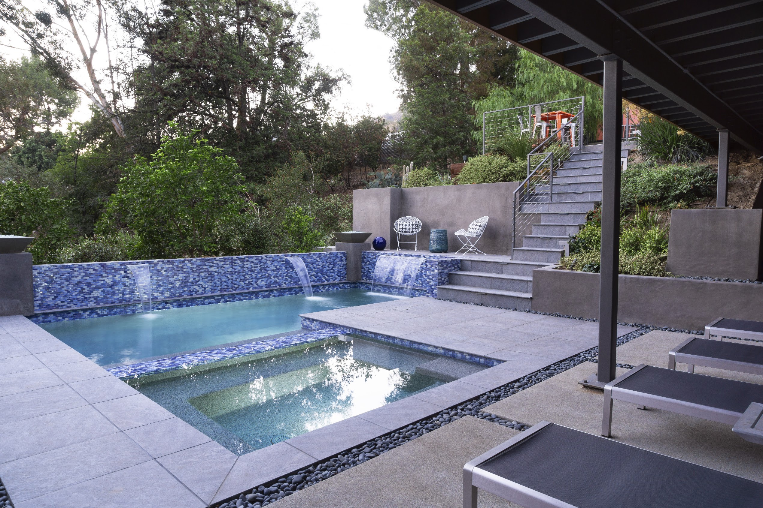 Serene modern backyard with a blue-tiled pool, raised spa, stone steps, lush landscaping, and shaded lounge chairs.