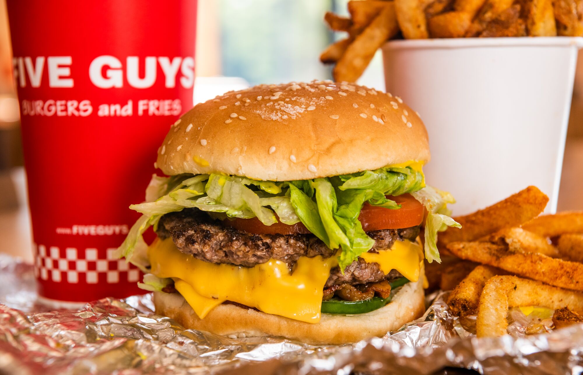 A close-up photograph of a Five Guys cheeseburger, soft drink in red Five Guys cup, and regular order of fries sitting on a table inside a Five Guys restaurant.