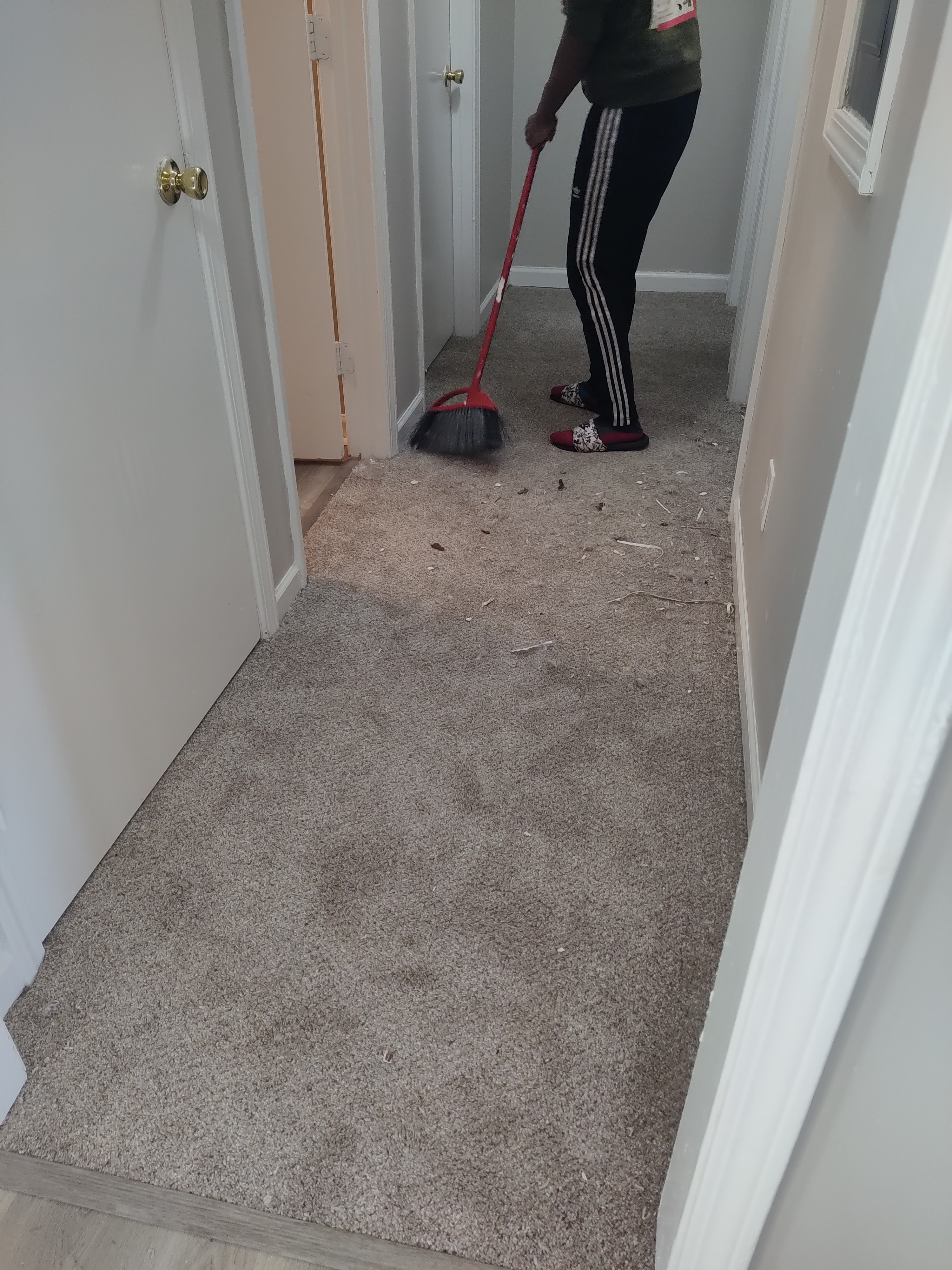 A person is sweeping debris off a carpeted hallway with a red broom. The person is wearing black athletic pants with white stripes and red slides. The hallway has several doors and light gray walls. The carpet is a neutral beige color.