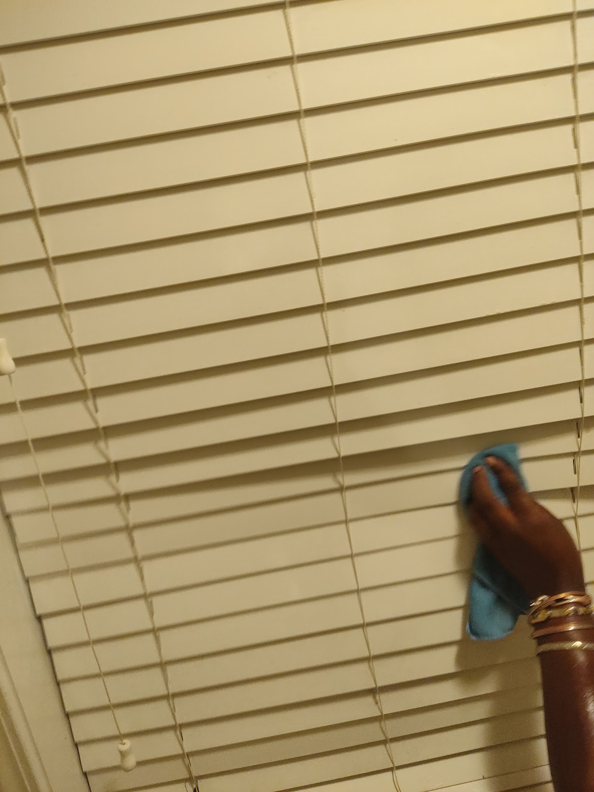 A person's hand, adorned with bangles, is wiping down a set of beige horizontal blinds with a blue cleaning cloth.