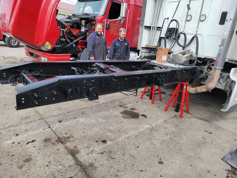 Two mechanics in dark blue work shirts stand behind a long, black semi-truck chassis frame that is e 