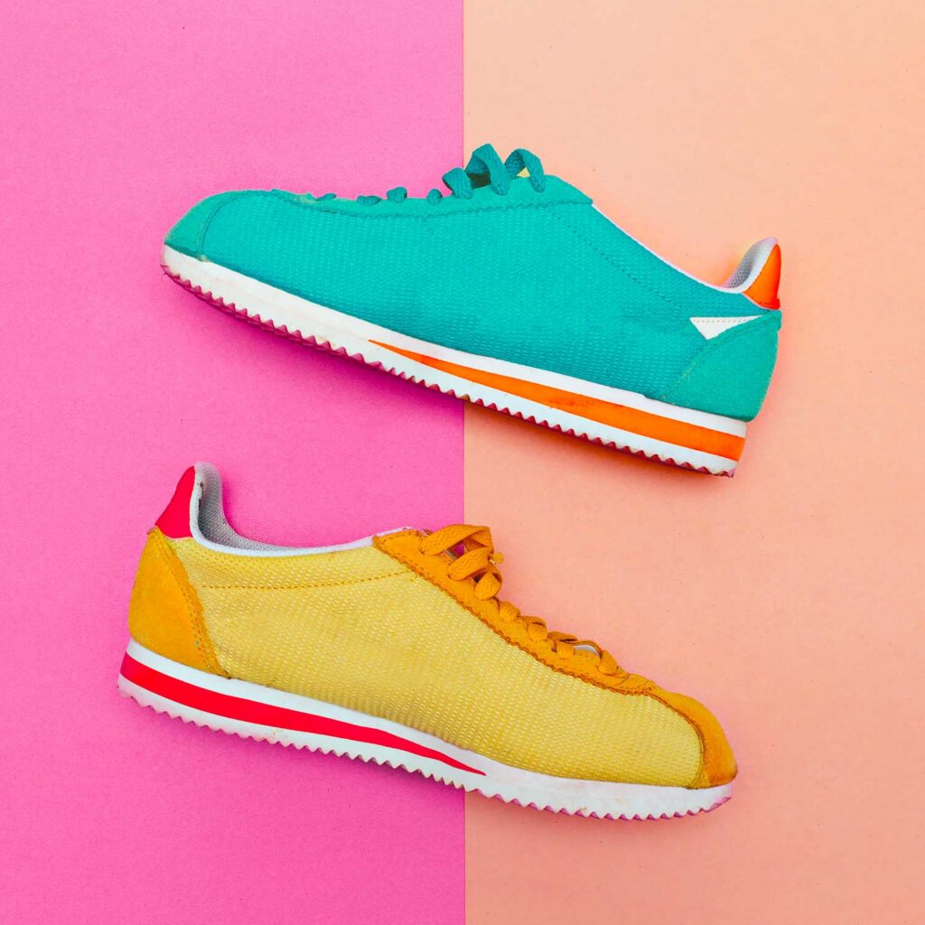 A pair of vintage-style athletic shoes, one teal and one yellow, are displayed on a split pink and peach background, creating a vibrant and retro aesthetic.