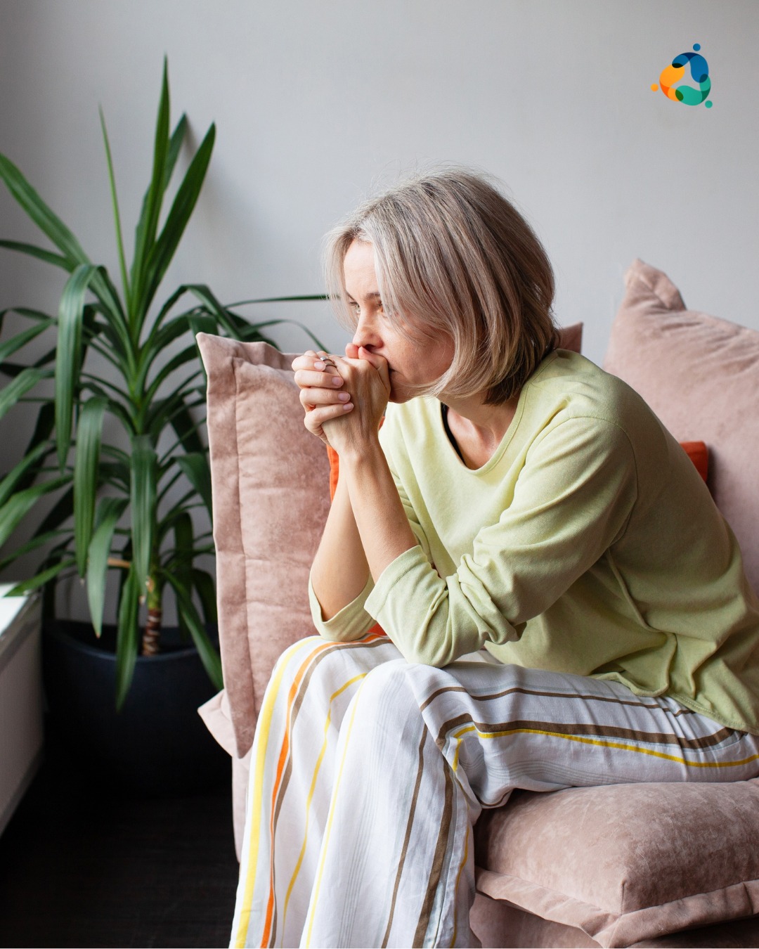 A woman sitting on a couch with her hands clasped and looking thoughtful or concerned, representing contemplation or emotional struggle.