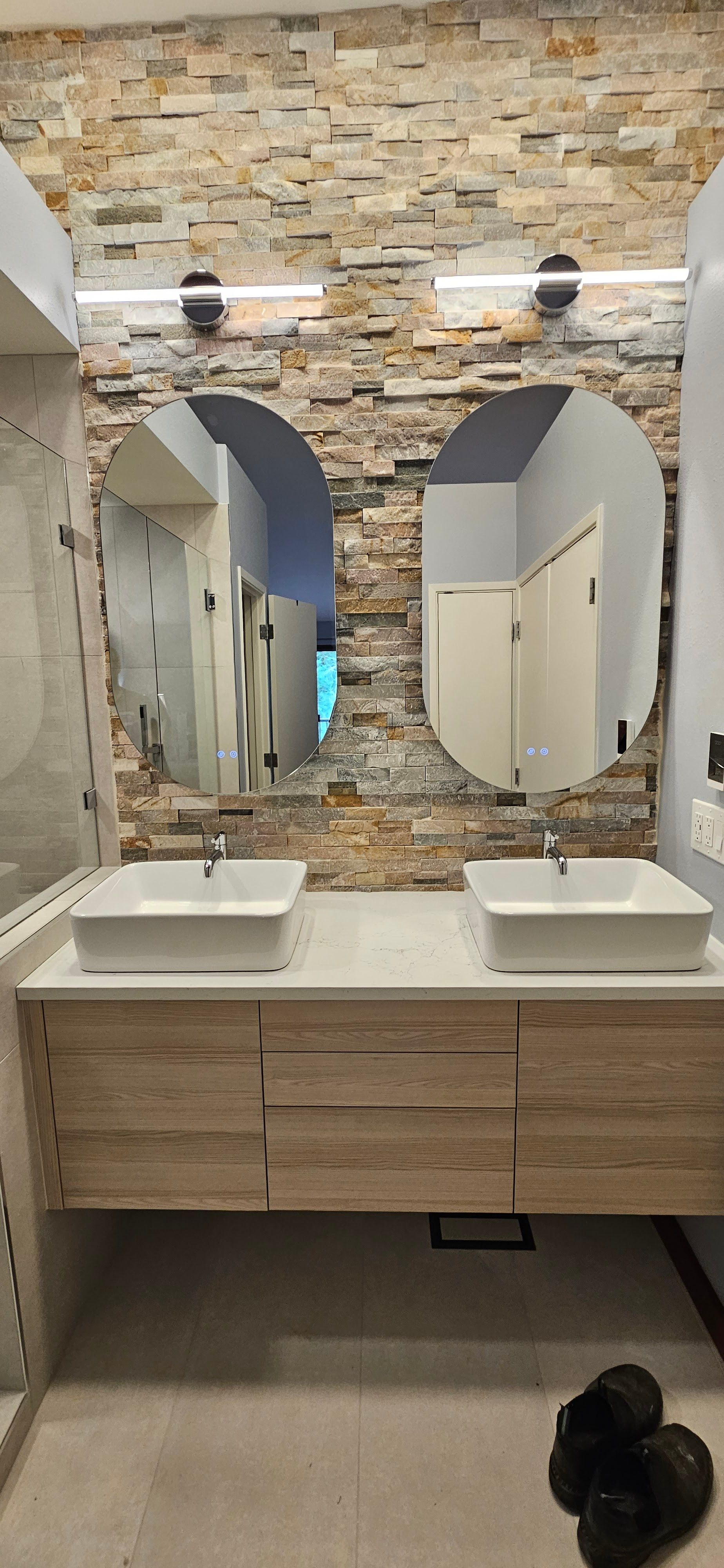 Modern double bathroom vanity featuring two white vessel sinks, oval mirrors, stone accent wall, and 