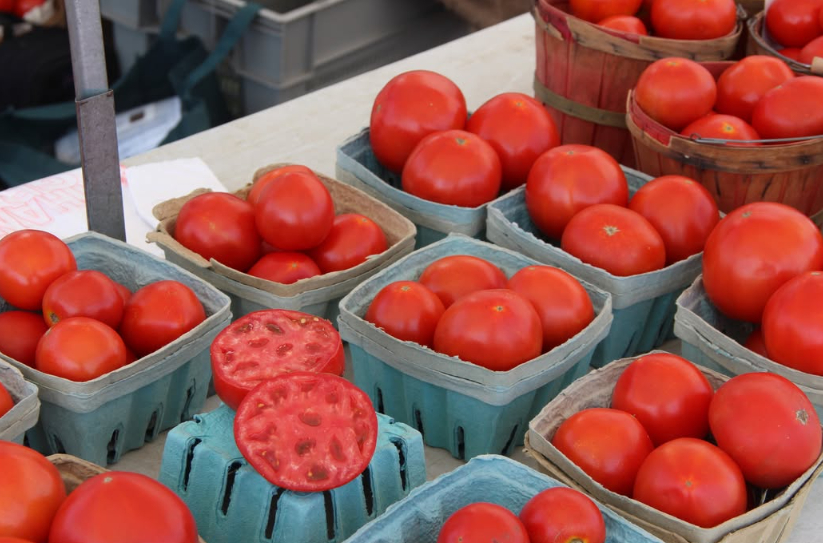 Containers and wooden baskets filled with smooth, ripe red tomatoes on a market stall table, featuring two sliced open halves highlighting juicy interiors.