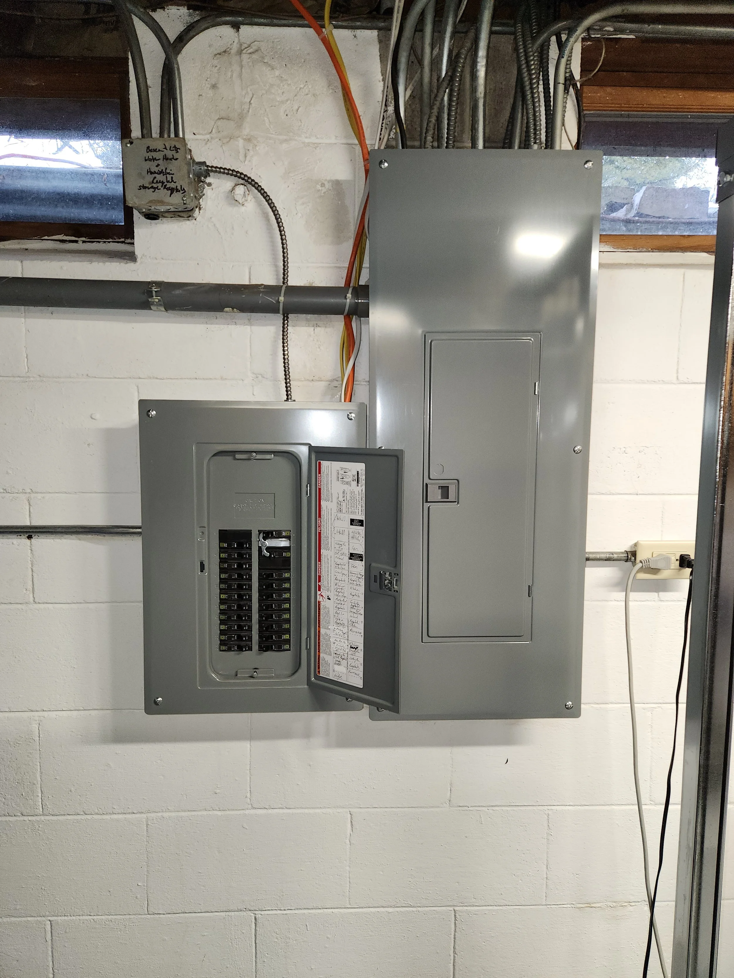 A close-up view of two electrical panels mounted on a white brick wall, with various electrical conduits and wires running across the top and connecting to the panels. One panel is open, revealing rows of circuit breakers, and the other panel is closed. An electrical outlet with two plugs is visible on the right side of the image, with cords plugged into it. A small, handwritten label is attached to a junction box near a window.