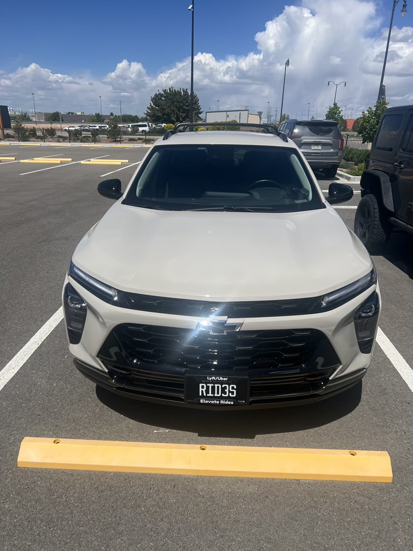 A white Chevrolet SUV is parked in a parking lot on a sunny day, with a yellow parking bumper in front of it. The car has a license plate that says "RID3S" and also mentions "Lyft/Uber" and "Elevate Rides". In the background, other cars and buildings are visible under a blue sky with scattered clouds.