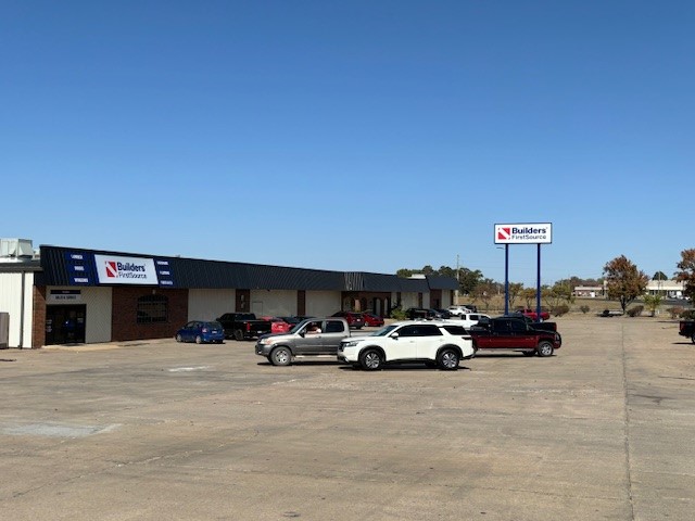 A wide shot of a Builders FirstSource facility with a parking lot in the foreground. The building has a mix of brick and metal siding with large signage displaying the Builders FirstSource logo above the entrance. Several cars and trucks are parked in the lot. To the right, a large standalone sign with the Builders FirstSource logo is visible near the street. The surrounding area includes trees and other commercial buildings under a clear blue sky, indicating a sunny day.