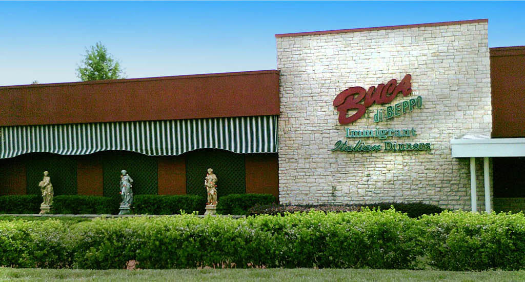 Buca sign on white brick with green and white striped cover area and greenery at Buca di Beppo Alpharetta.