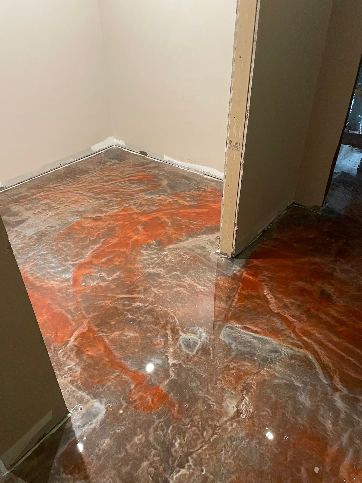 A close-up, low-angle shot of a newly installed epoxy floor with a metallic, marbled effect in shades of orange, brown, and white. The floor has a high-gloss, reflective surface, showing highlights from overhead lighting. The room appears to be in a state of renovation, with unfinished drywall visible at the corners and around a doorway.