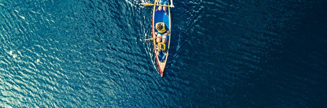 Aerial view of a small, colorful fishing boat with two people, surrounded by vast, rippling blue water. The scene conveys peacefulness and solitude.