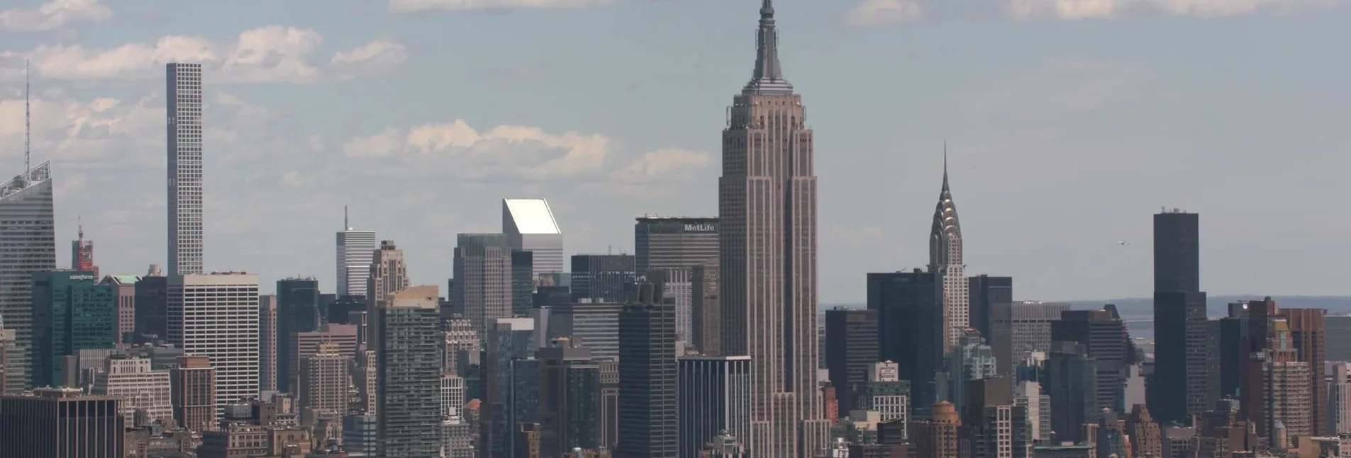 A panoramic view of the New York City skyline on a partly cloudy day, featuring iconic skyscrapers such as the Empire State Building and the Chrysler Building, as well as modern architectural marvels like 432 Park Avenue. The scene captures the density and verticality of Manhattan's urban landscape.