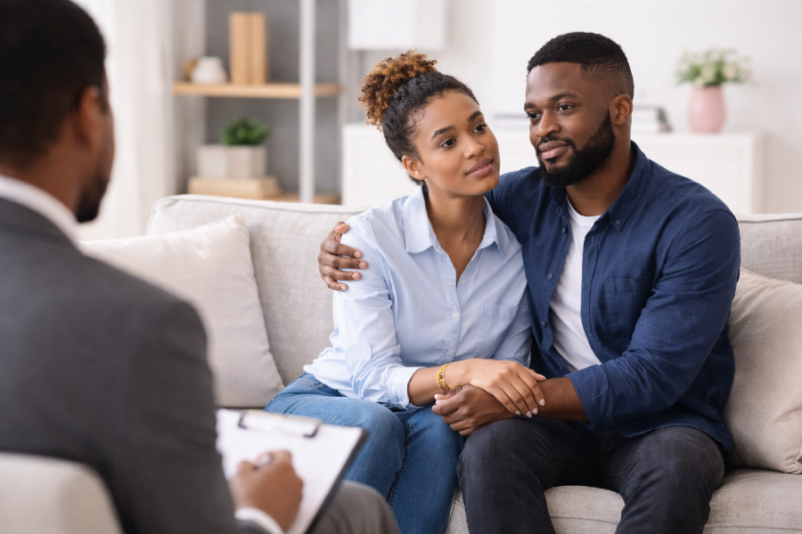 A couple sits close together on a couch, holding hands, while a person in a suit takes notes in the foreground, suggesting a therapy or counseling session.