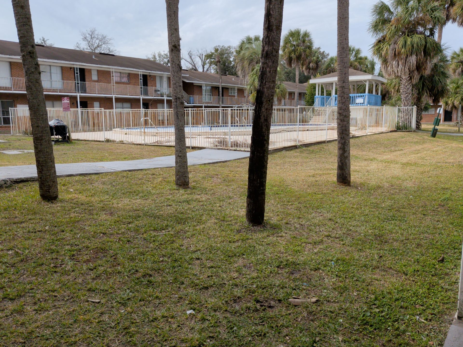 Apartment complex outdoor area with grassy lawn, palm trees, and fenced swimming pool. Two-story brick residential buildings surround the community pool and gazebo seating area.