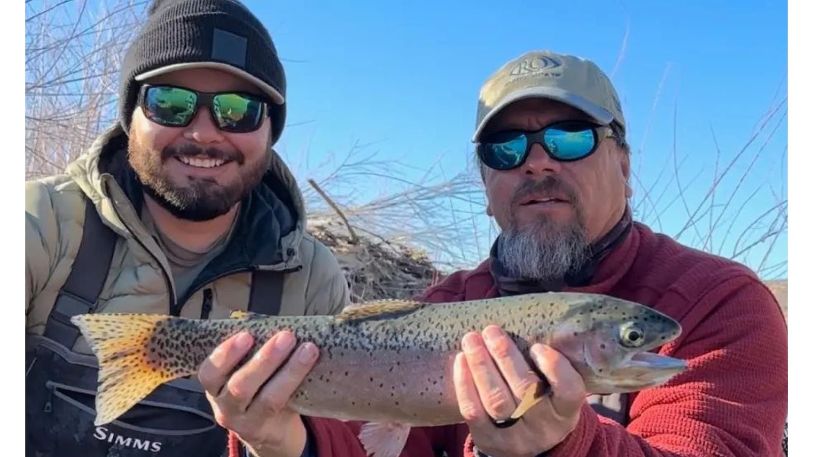 Two men are holding a freshly caught rainbow trout outdoors, with a clear blue sky in the background. Both men are wearing fishing attire, including hats and sunglasses, and appear to be smiling. The trout is colorful with spots and has its mouth slightly open. The man on the left is wearing a black beanie and a green puffer vest, while the man on the right is wearing a baseball cap and a red long-sleeved shirt. The lower portion of the image shows the brand name "SIMMS" on the fishing waders worn by the man on the left.