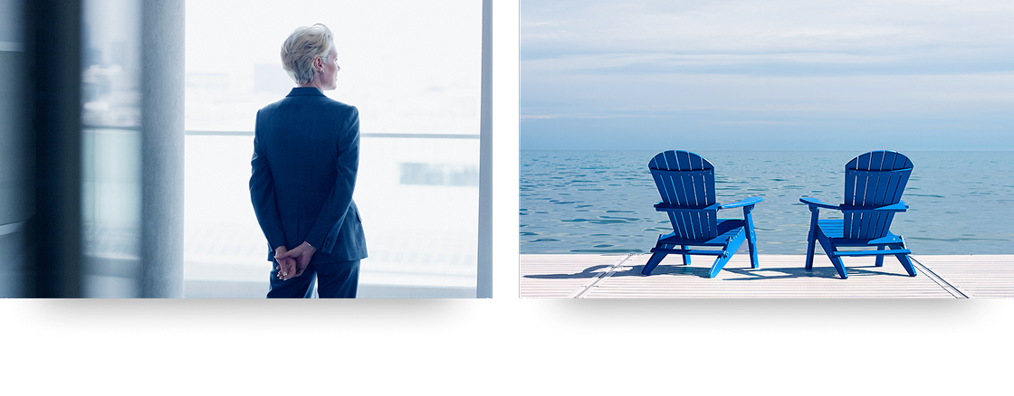 Short-haired woman wearing a suit staring out a large window with her hands crossed behind her back & two blue Muskoka chairs on a dock facing out towards the lake.