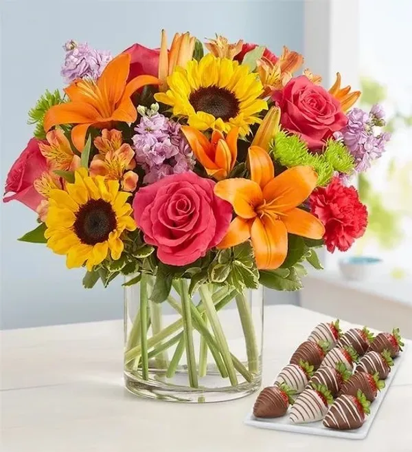 A vibrant bouquet of sunflowers, lilies, roses, and carnations in a clear glass vase sits on a white 