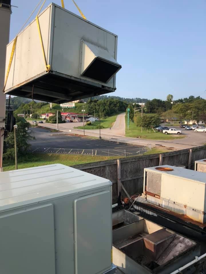 Rooftop HVAC unit being lifted by crane using straps, positioned above building during installation or replacement, with surrounding road and landscape visible, demonstrating commercial HVAC service and equipment handling operation.