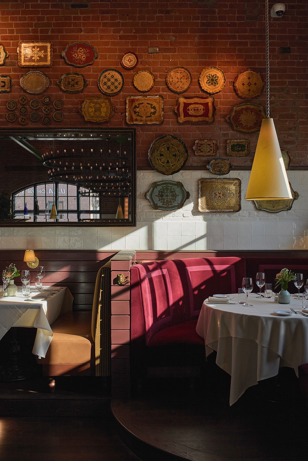 A decorative wall in a restaurant featuring dozens of antique, colorful serving trays mounted on exposed brick. Below, a plush red curved booth is illuminated by a streak of sunlight.