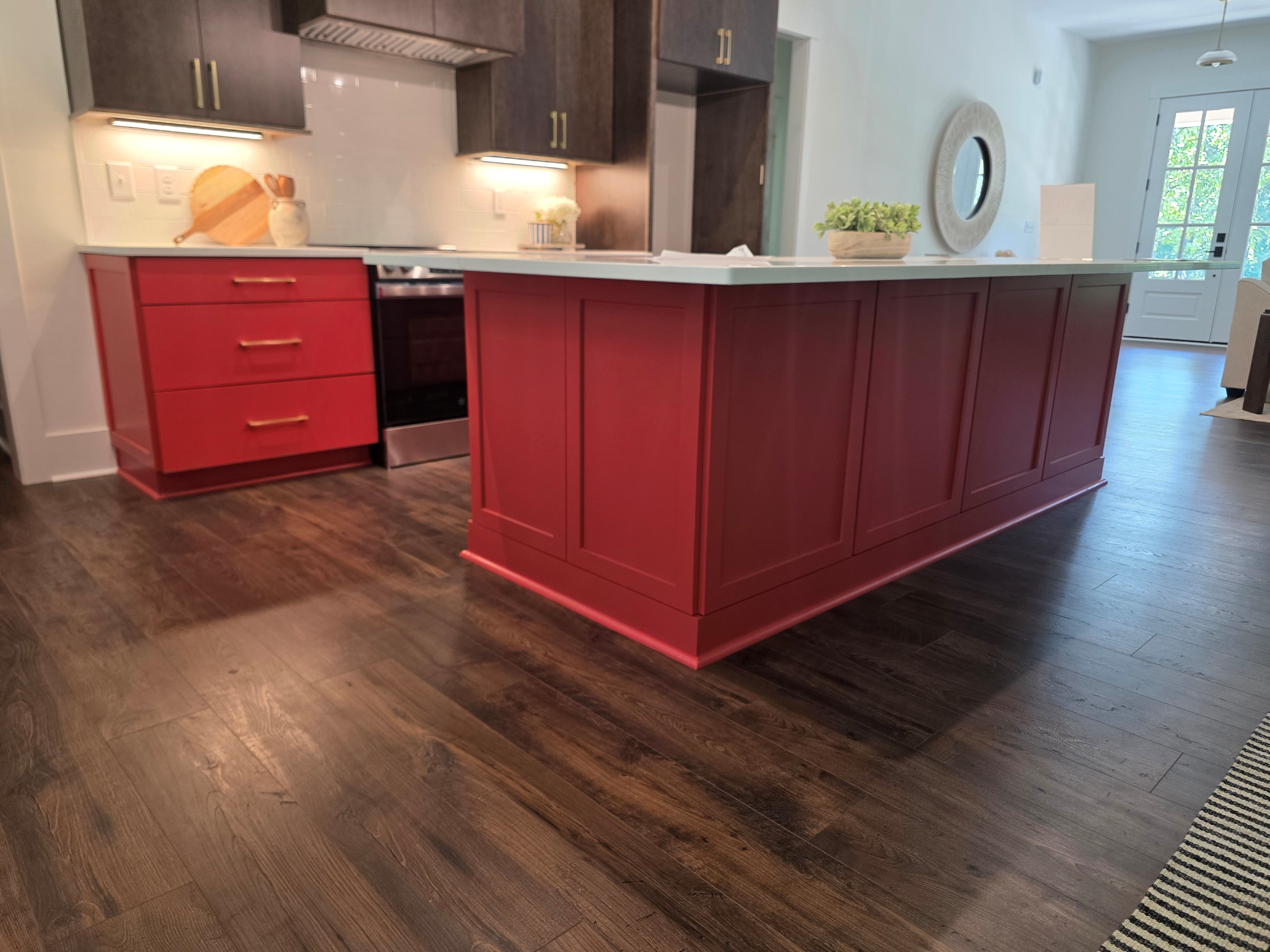 Kitchen view highlighting a large red island with paneled cabinetry and white countertop, complemented by dark wood floors and surrounding red cabinetry.