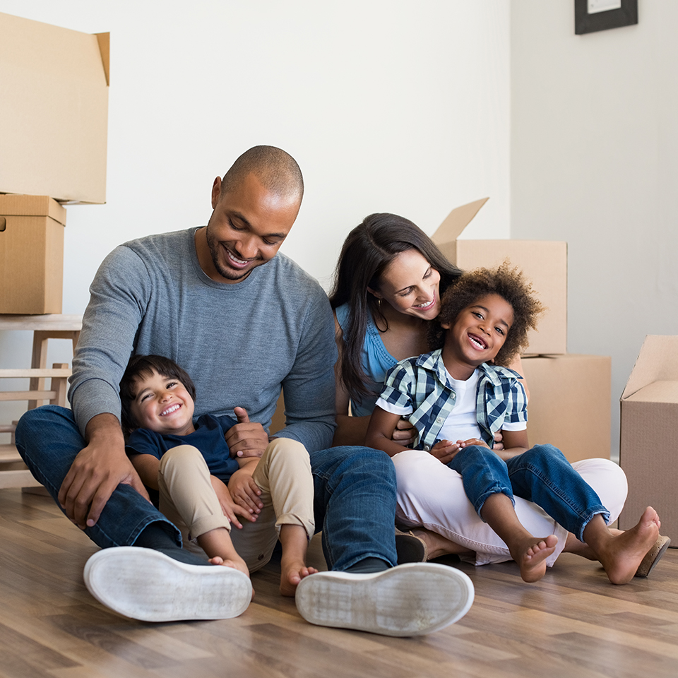 A man and woman and two kids sitting on hardwood floors surrounded by cardboard boxes.
