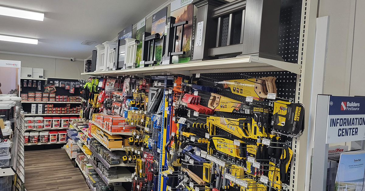 Interior aisle at a Builders FirstSource showroom displaying Stanley hand tools, saws, and tape measures on pegboard hooks, with door and window sample displays mounted above. A Builders FirstSource Information Center sign is visible at the end of the aisle.