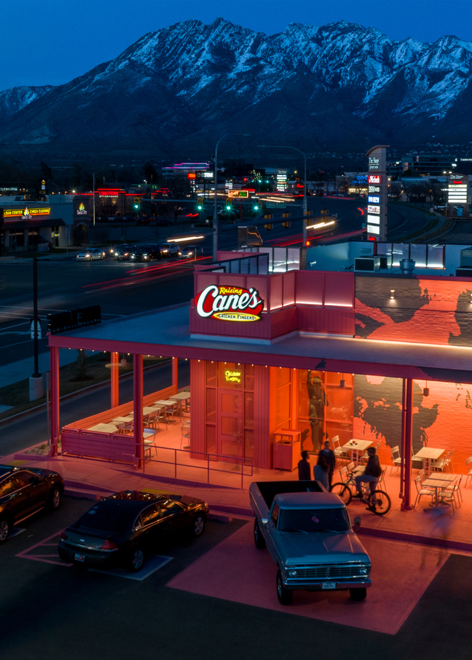 A custom, bright pink Raising Cane's restaurant glowing at twilight, set against a dramatic backdrop of massive snow-capped mountains. The building features a large outdoor patio where people are gathering, and a vintage silver pickup truck is parked in the foreground of the pink parking lot.