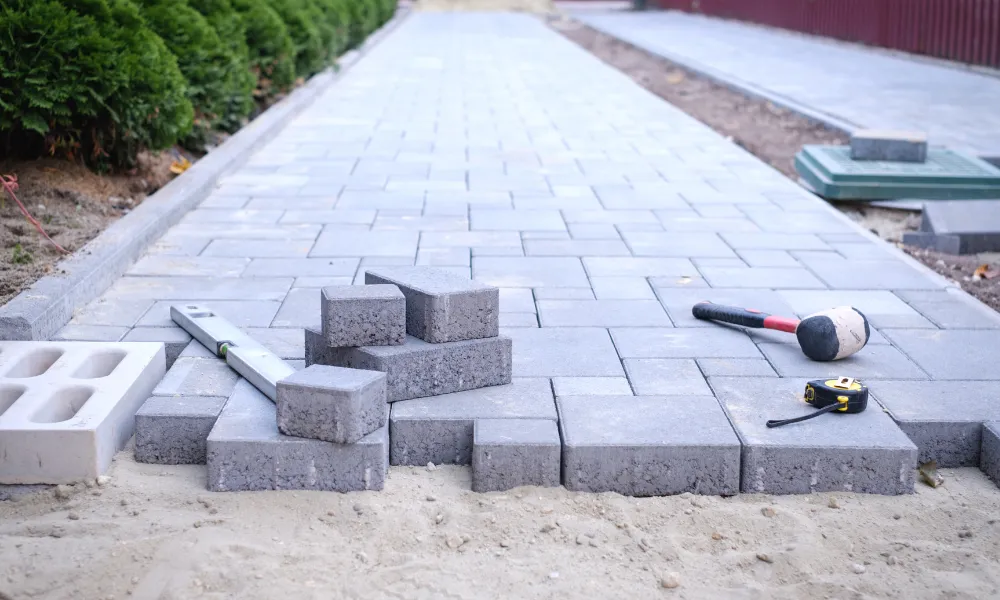 A sidewalk is under construction, with grey paving stones laid out in a pattern on a sand base. Tools such as a level, rubber mallet, and measuring tape are placed on the stones, indicating active work. A row of green bushes lines the left side of the walkway, and a reddish-brown fence is visible in the background to the right.