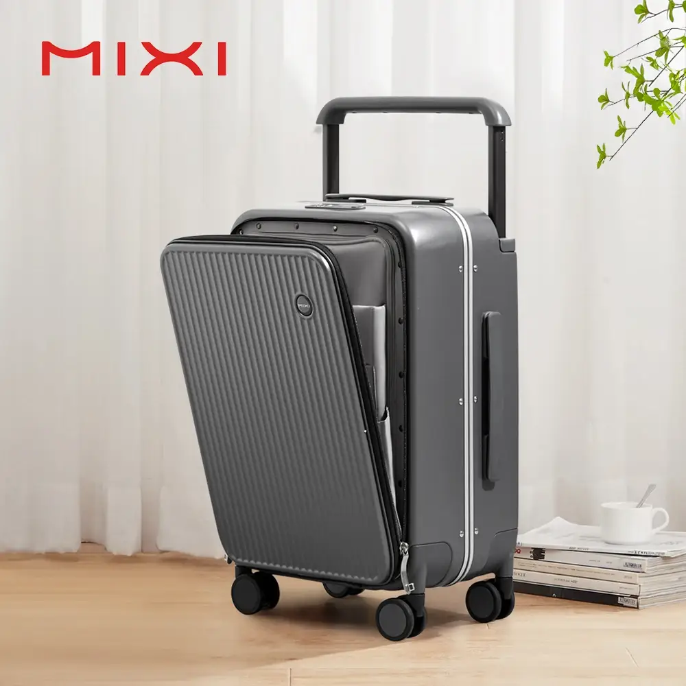 A sleek, dark gray suitcase stands open, revealing its interior. The suitcase has a ribbed textured front panel that is partially open, showing neatly organized compartments. It is equipped with four spinner wheels and a retractable handle, suggesting it's designed for travel. The background features sheer white curtains and a stack of books with a white cup and saucer on top. A small sprig of green leaves is visible in the upper right corner. The brand logo 'MIXI' is prominently displayed in red at the top left of the image.